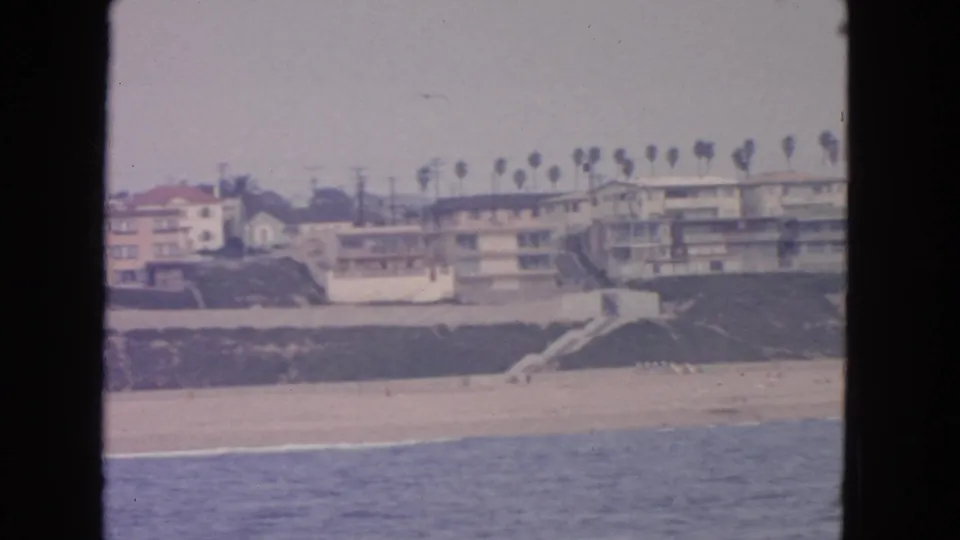 Offshore 8mm view of a California shoreline with beach, bluff, palms, and low residential buildings, dated 1963 by clip metadata.