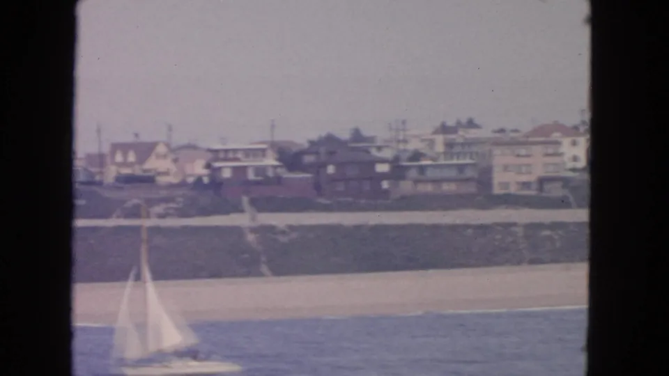 Small sailboat crossing the foreground with a sandy beach, grassy bluff, and shoreline houses behind it in 1963 coastal footage.