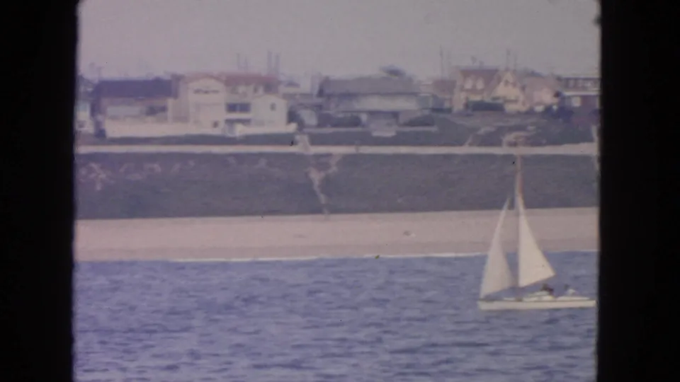 1963 8mm frame of a small sailboat offshore, with a quiet beach, bluff, and coastal homes behind it in San Francisco metadata context.