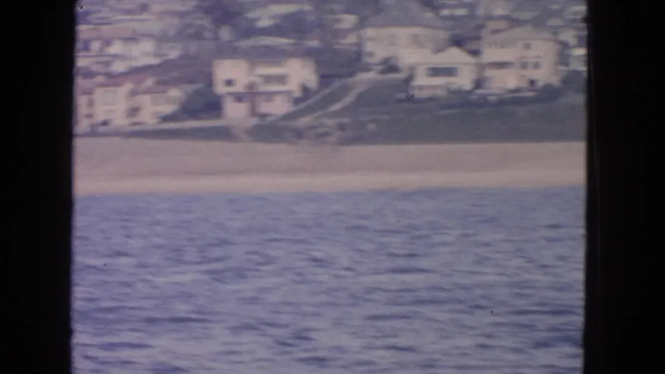 Vintage 8mm frame of the San Francisco beach from offshore, showing sand, water, homes, and a hillside path.