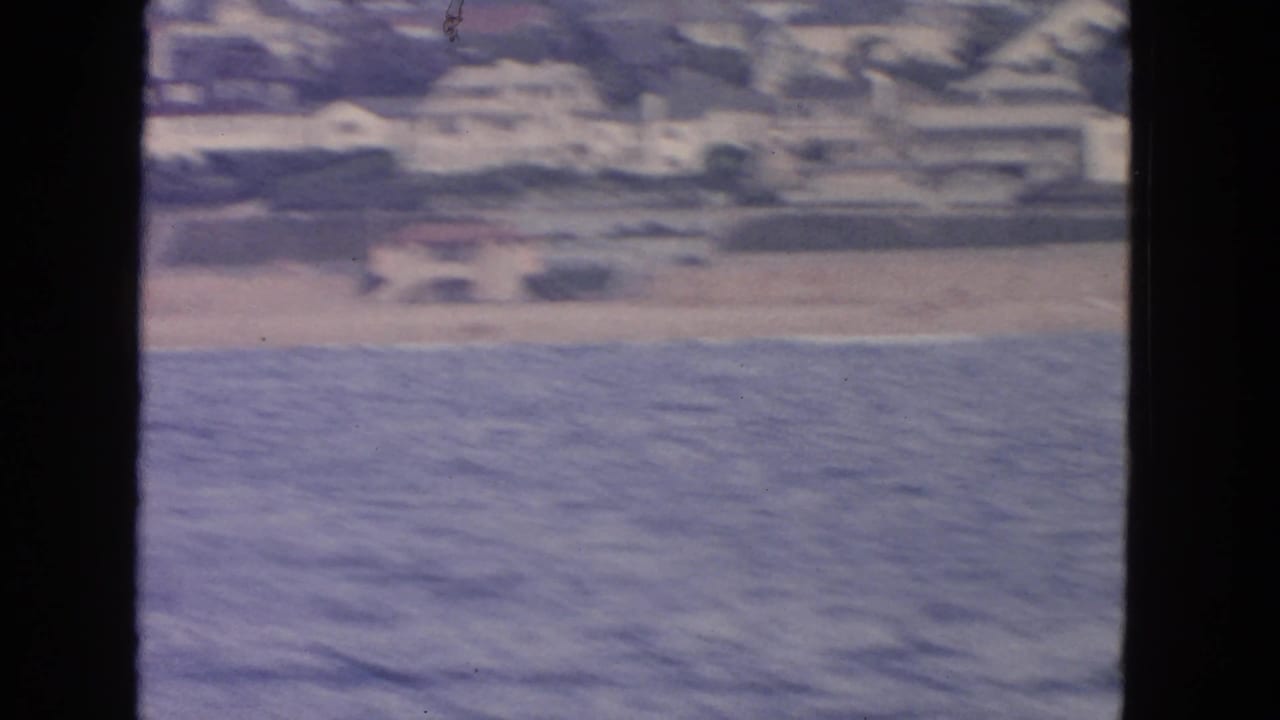 1963 San Francisco home-movie frame from a boat, showing blue water, a beach strip, and soft-focus shoreline buildings.