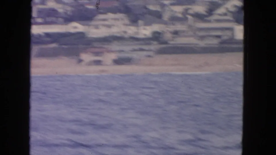1963 San Francisco home-movie frame from a boat, showing blue water, a beach strip, and soft-focus shoreline buildings.