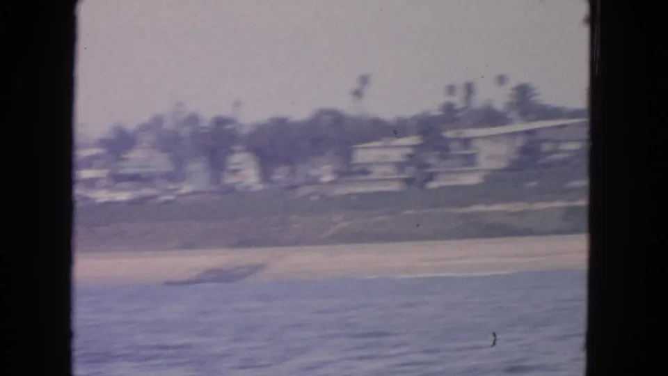 Soft-focus 1963 coastal frame showing water, a pale beach edge, and distant shoreline structures from a moving boat.