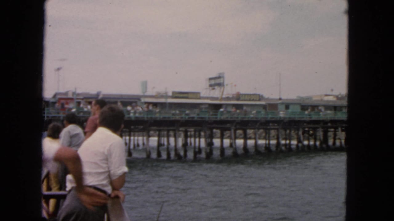 Passengers stand near a boat railing as the pier and waterfront signs pass behind them.