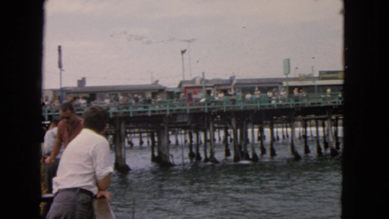 1963 San Francisco home-movie frame with boat passengers looking toward a busy wooden pier with green railings and pilings.
