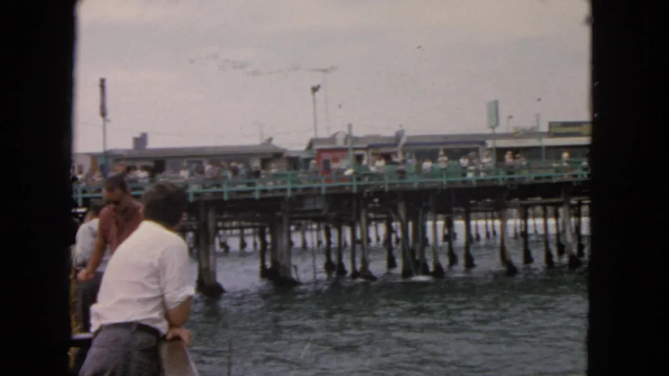 1963 San Francisco home-movie frame with boat passengers looking toward a busy wooden pier with green railings and pilings.