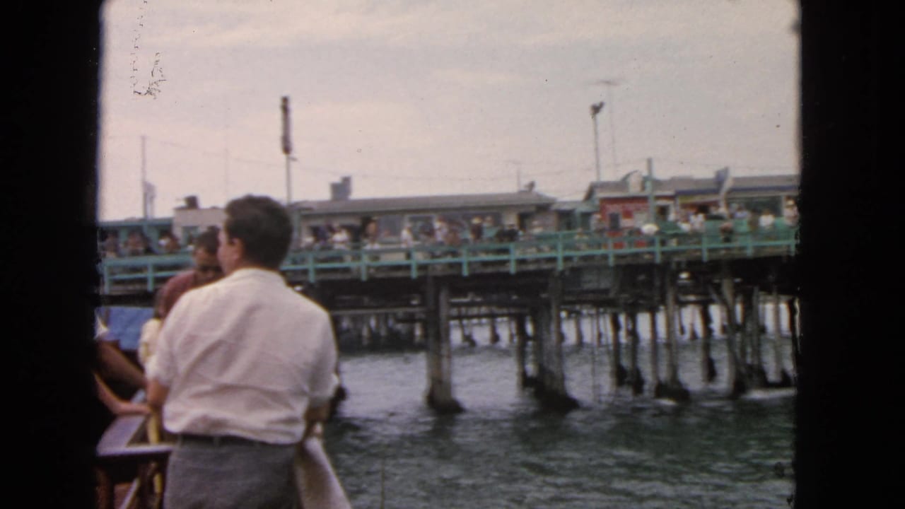 People on a boat stand near the rail while a busy pier fills the background.