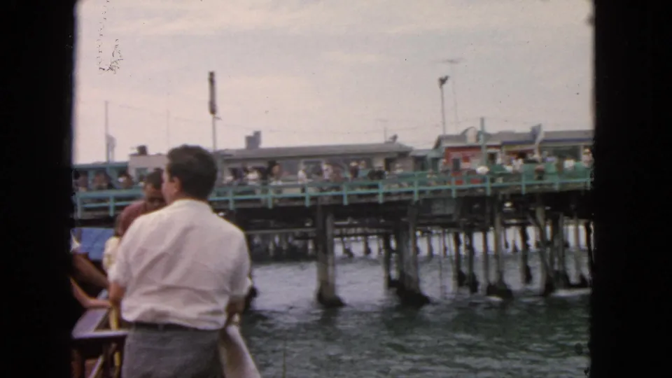 Boat passengers in 1963 San Francisco watch a crowded waterfront pier with green railings, pilings, and small buildings.