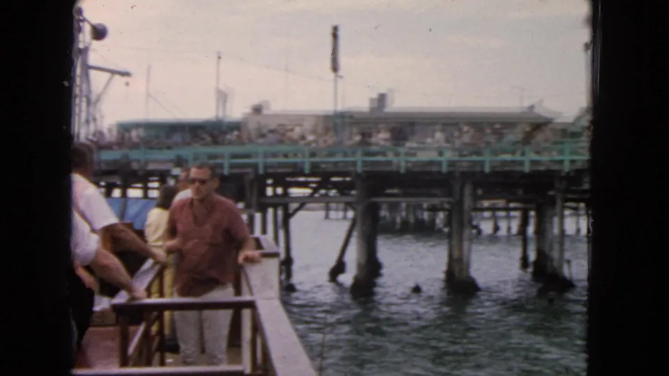 A man in sunglasses is seen near the boat rail as the 1963 San Francisco pier and waterfront crowd pass in the background.