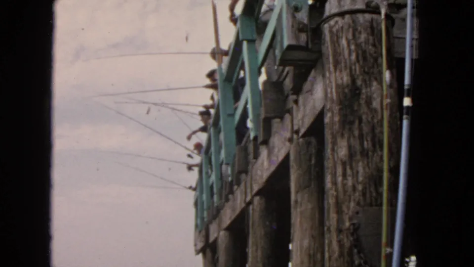1963 San Francisco waterfront frame showing the side of a wooden pier, green railings, and fishing rods projecting outward.