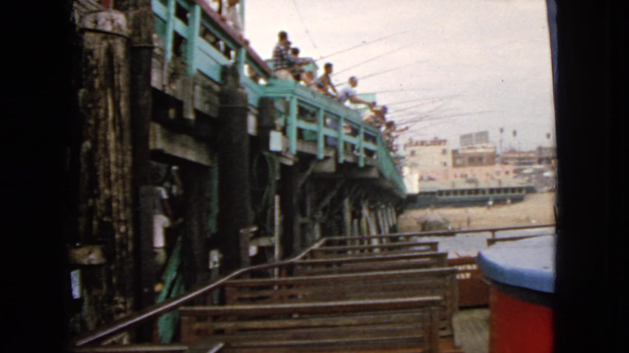 1963 San Francisco home-movie frame showing people on a green-painted ocean pier, with pilings, railings, and water below.