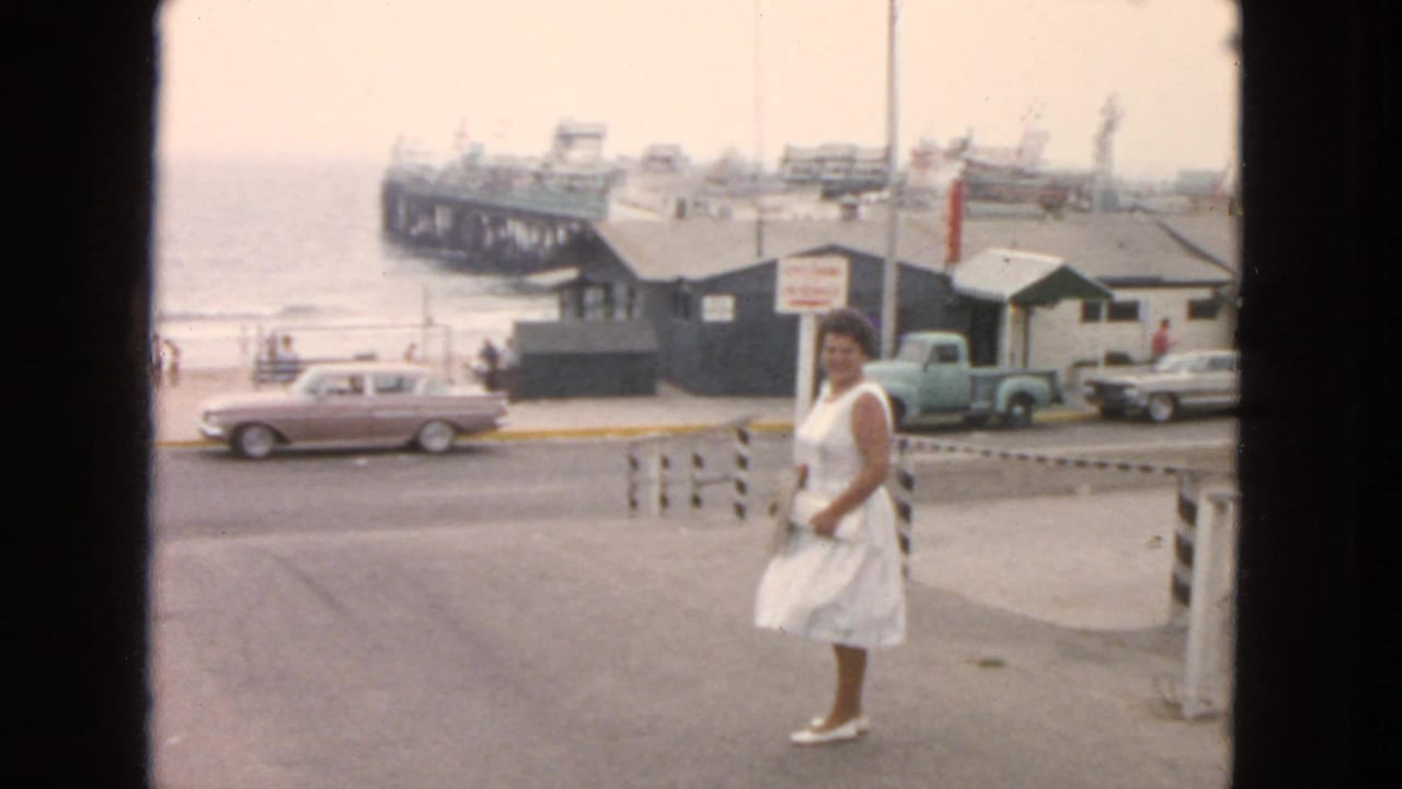 1963 San Francisco waterfront view with a woman in a white dress, older cars, pier buildings, and hazy water in the background.