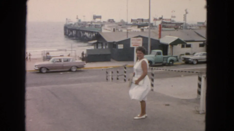 Home-movie frame from the San Francisco waterfront showing a woman walking near older cars, low pier buildings, and the shoreline.