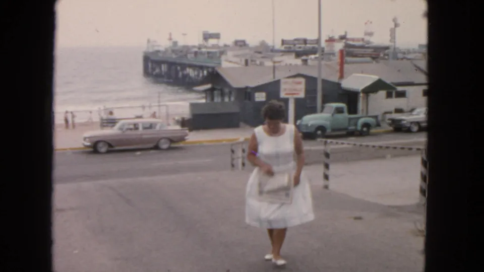 1963 San Francisco waterfront frame showing a woman in a white dress carrying a small bag, with cars, pier buildings, and ocean behind her.