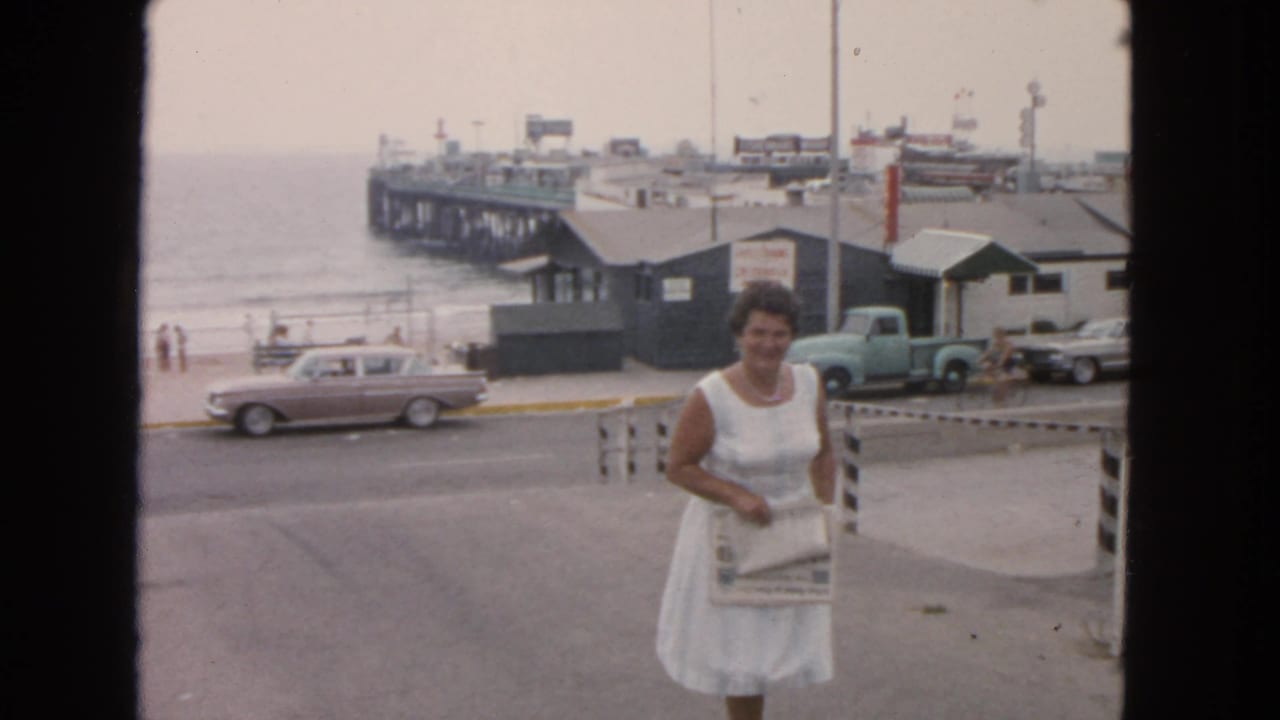 In 1963 San Francisco, a woman in a white dress walks uphill holding a paper, with a pier, beach, vintage cars, and a truck in the background.