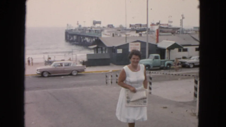 In 1963 San Francisco, a woman in a white dress walks uphill holding a paper, with a pier, beach, vintage cars, and a truck in the background.
