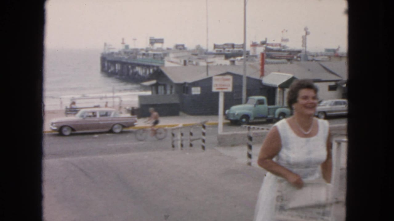 Woman exits frame at right as a cyclist passes along the waterfront road near parked cars and a pier.