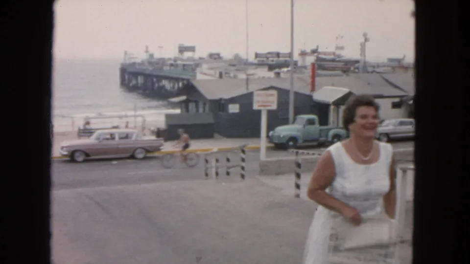 In the San Francisco waterfront scene, a woman in white moves out of frame while a cyclist rides past mid-century cars, beach buildings, and a pier.
