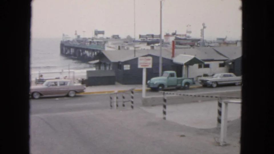 The woman is no longer visible as the frame holds on a 1963 San Francisco waterfront road with vintage cars, a light blue pickup, beach, and pier.