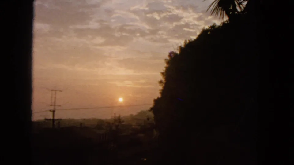 1963 8mm San Francisco dusk view with the sun in a hazy sky, dark hillside vegetation, silhouetted wires, and soft film exposure.