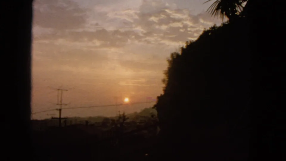 1963 San Francisco 8mm frame showing a low orange sun, silhouetted hillside, utility poles, wires, and softly clouded dusk sky.
