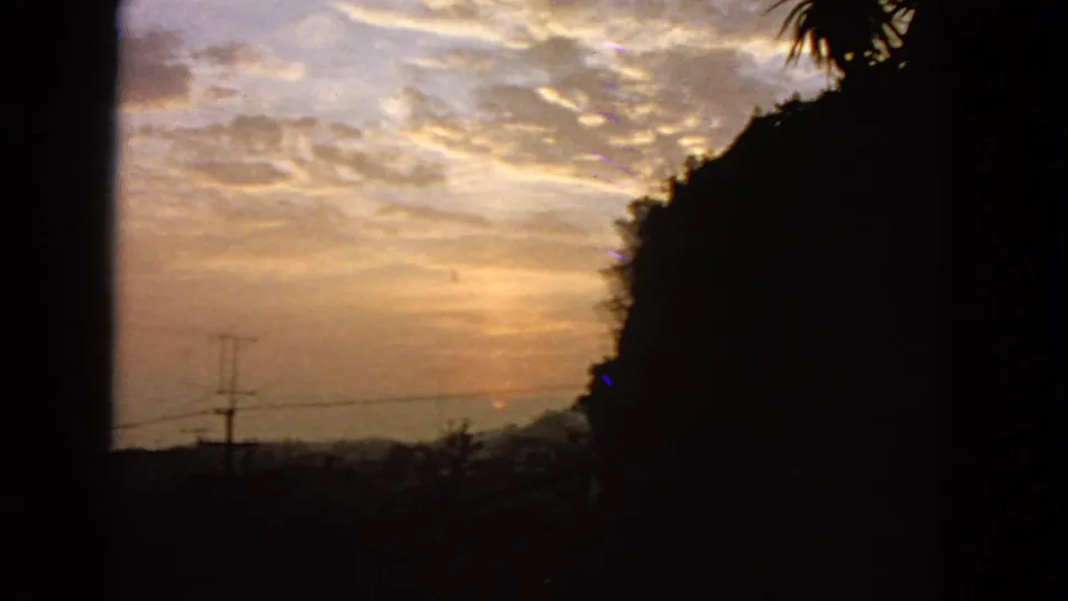 1963 San Francisco home-movie frame of the sun nearly down, with utility wires, dark hillside mass, palm fronds, and clouded dusk sky.