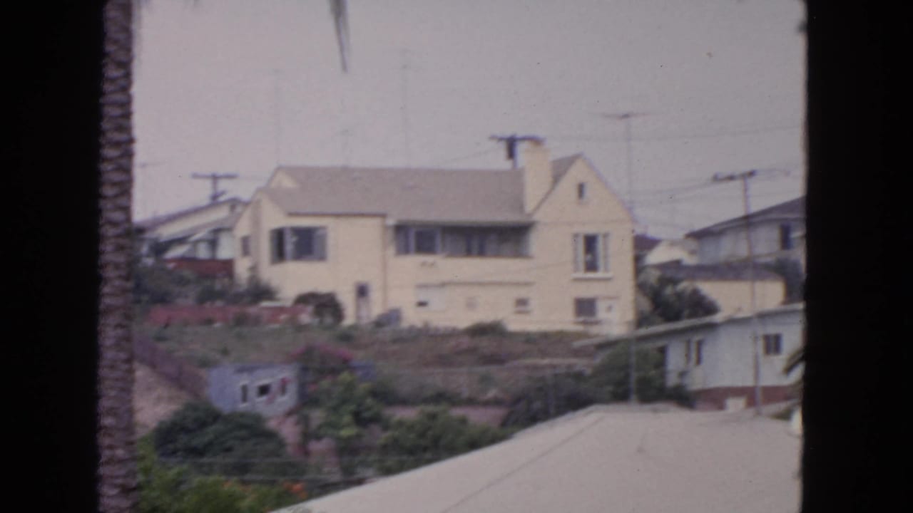 Yellow two-story hillside house seen past a palm trunk and nearby rooftops.