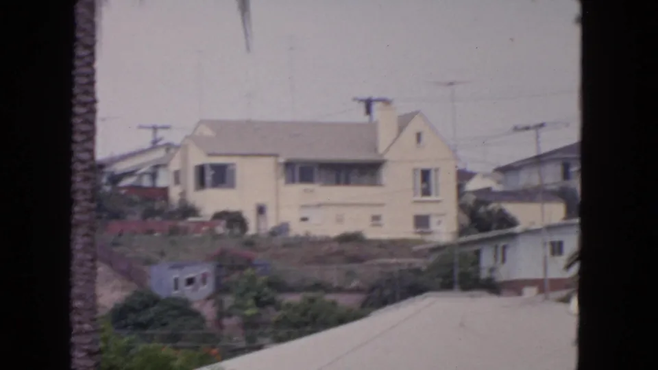 1963 San Francisco hillside home view with a pale yellow two-story house, palm trunk, utility poles, and surrounding greenery.