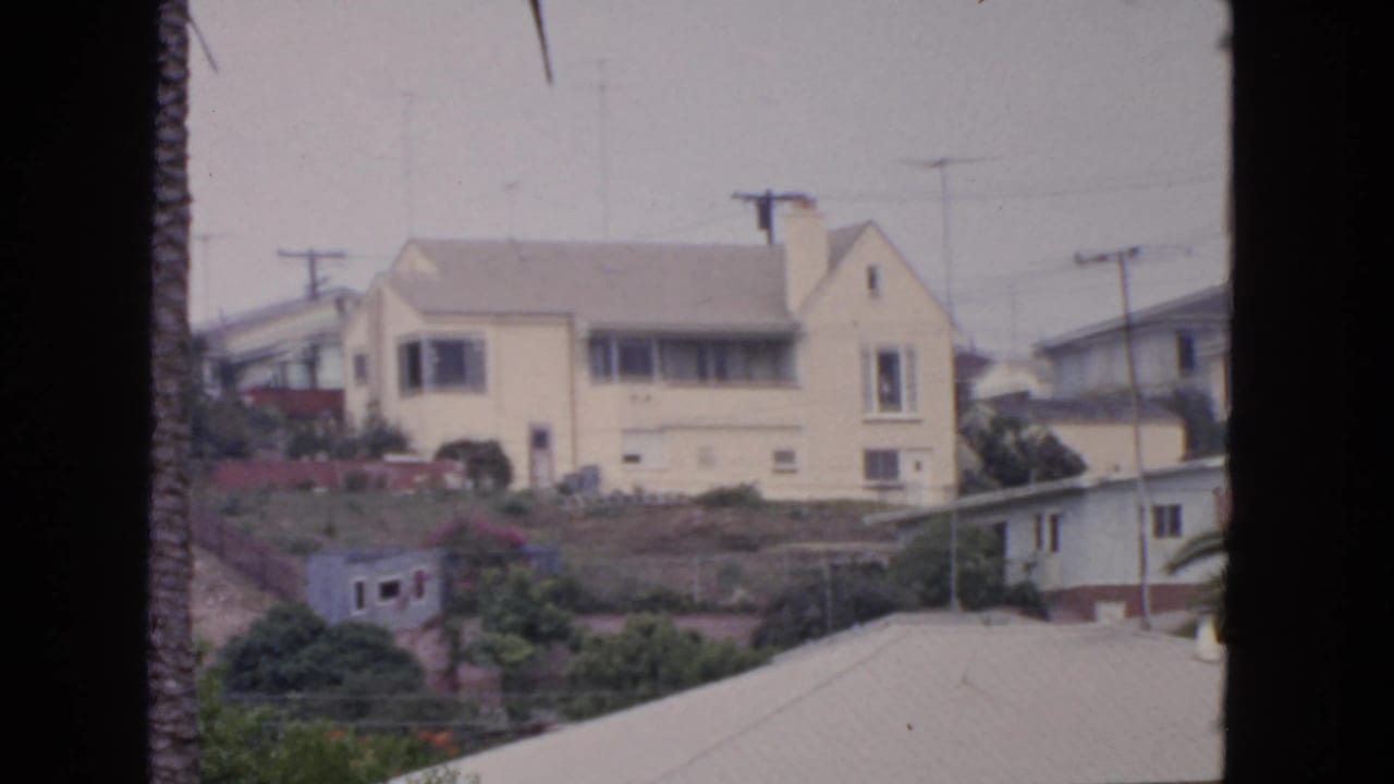 Hillside house with pale exterior, antennas, utility poles, and garden plantings.