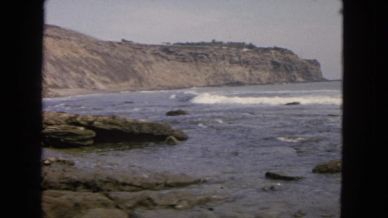 Rocky coast with waves breaking below distant cliffs, framed by dark vertical edges.