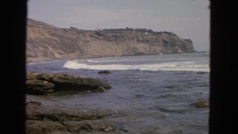 1963 San Francisco shoreline footage showing low surf, scattered rocks, and pale coastal cliffs through a dark foreground frame.