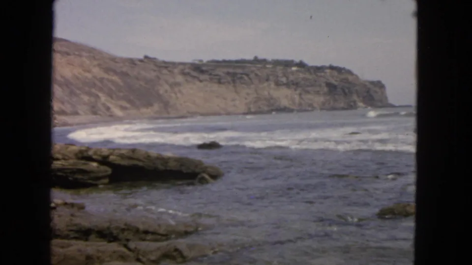 8mm 1963 San Francisco coastal scene with foamy surf, weathered rocks, and a distant bluff under a muted sky.