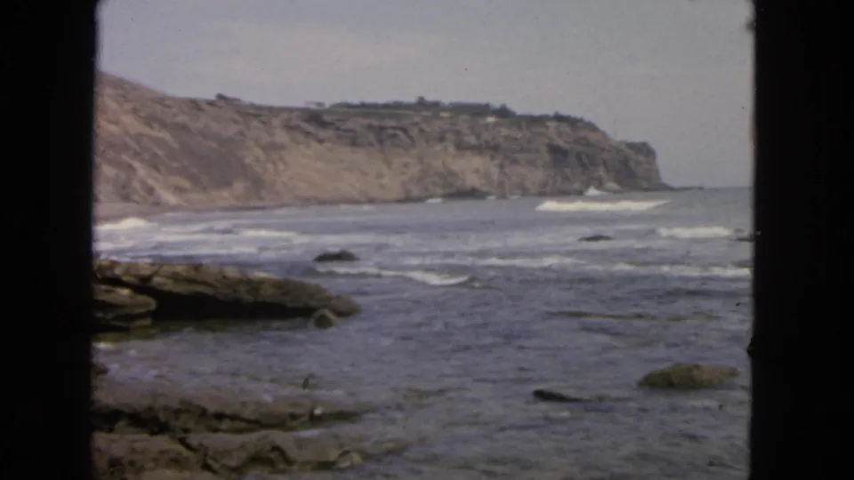 1963 San Francisco 8mm view of waves rolling over coastal rocks, with distant cliffs and dark vertical borders in the foreground.