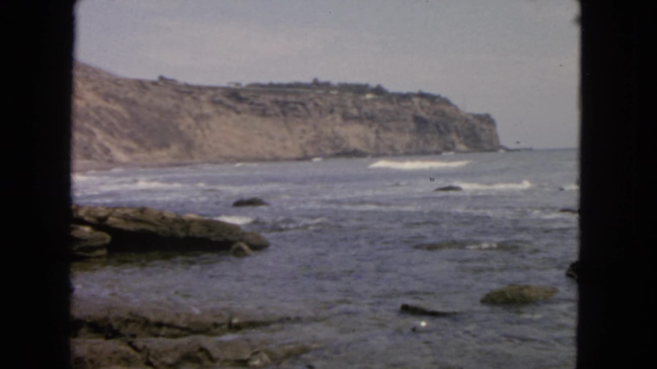 1963 San Francisco coastal view with waves breaking over rocks below a distant cliff, framed by dark window or doorway edges.