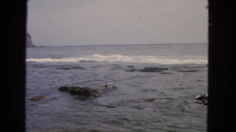 1963 San Francisco coastal view with shallow surf, exposed rocks, and a partial distant cliff under muted overcast light.