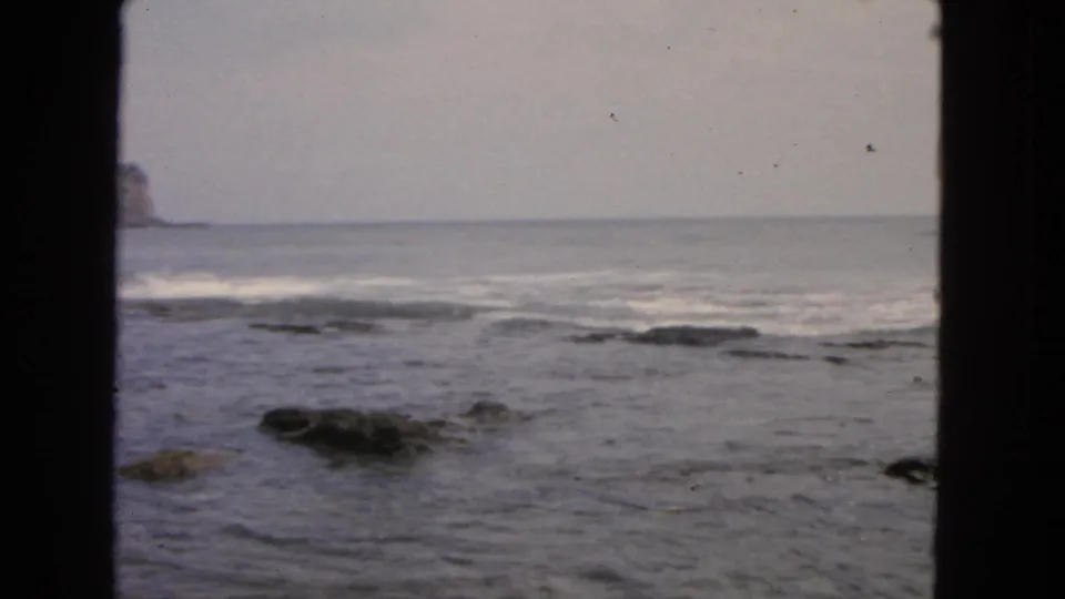 1963 San Francisco ocean cutaway showing dark rocks in shallow water, soft surf, and a pale overcast horizon.
