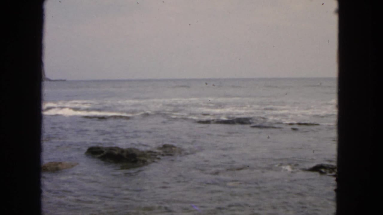 Ocean waves breaking over low shoreline rocks under a bright hazy sky.