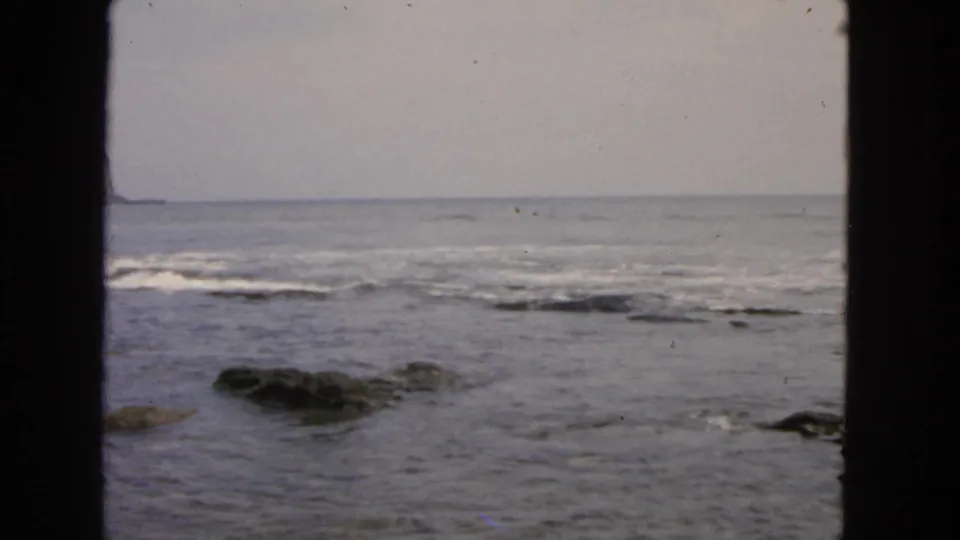 1963 San Francisco coastal home-movie frame showing surf and low rocks, bordered by dark 8mm scan edges.