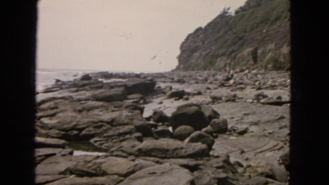 1963 San Francisco shoreline view with layered rocks, rounded boulders, surf, and a grassy cliff in faded 8mm color.