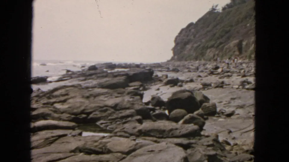 Home-movie frame from 1963 San Francisco showing a rocky shoreline, distant surf, and cliff face with dark 8mm frame edges.