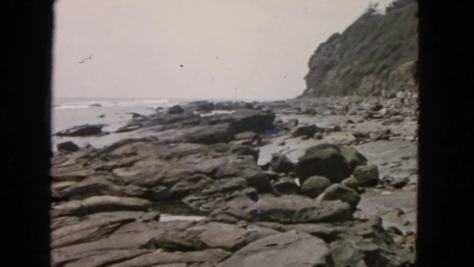 1963 San Francisco coastal frame showing shallow surf around dark rocks, rounded boulders, and a cliff in soft 8mm color.