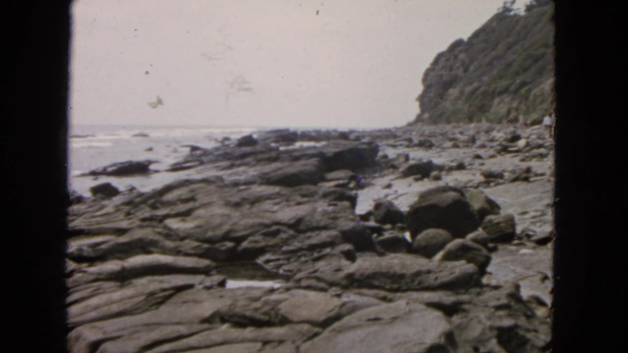 1963 San Francisco coastal home-movie frame showing layered rocks, boulders, ocean waves, and a vegetated cliff under a pale sky.