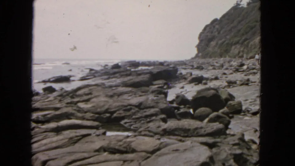 1963 San Francisco coastal home-movie frame showing layered rocks, boulders, ocean waves, and a vegetated cliff under a pale sky.