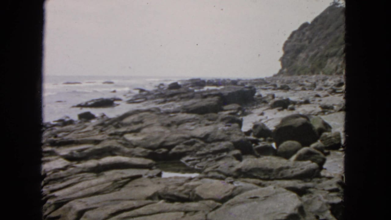 1963 San Francisco shoreline view with dark film edges, flat rock shelves, scattered boulders, gentle surf, and a cliff rising at right.
