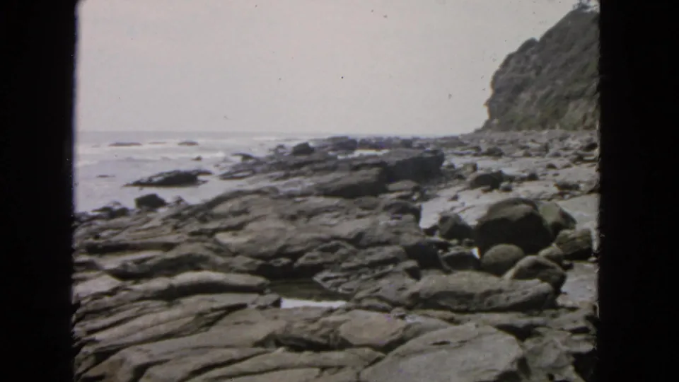 1963 San Francisco shoreline view with dark film edges, flat rock shelves, scattered boulders, gentle surf, and a cliff rising at right.