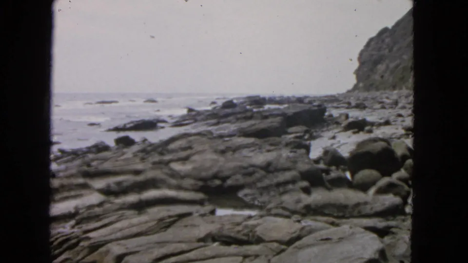 1963 California coastal frame showing low surf, wet rock shelves, rounded stones, and a steep vegetated cliff along the right edge.