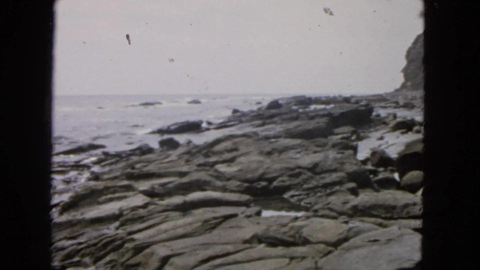 1963 San Francisco coastal home-movie frame with layered shoreline rocks, small waves, a pale ocean horizon, and cliff vegetation at right.