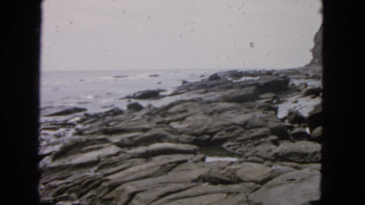 1963 San Francisco 8mm frame showing layered rocks beside the ocean under an overcast sky.