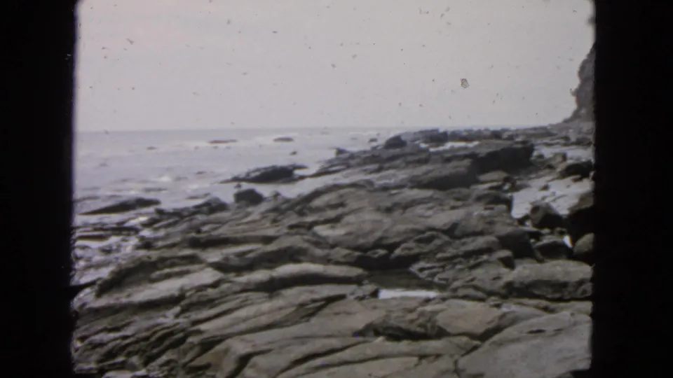 1963 San Francisco 8mm frame showing layered rocks beside the ocean under an overcast sky.