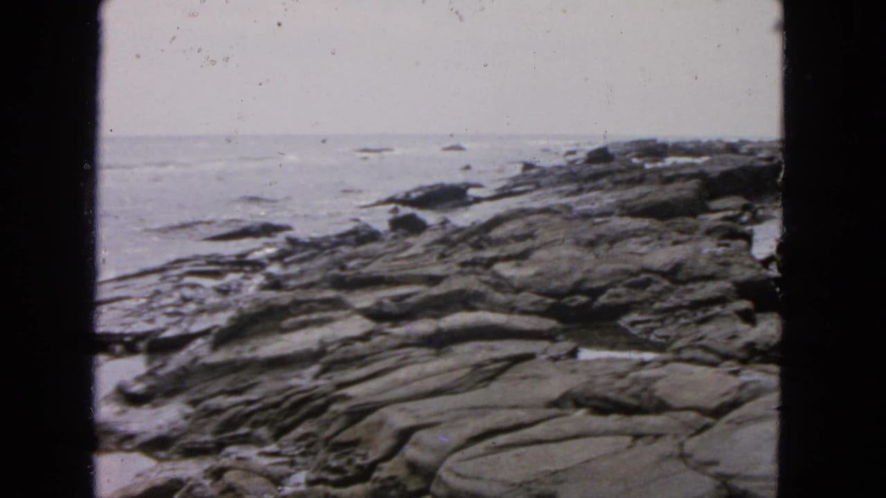 1963 San Francisco coastal home-movie frame with dark shoreline rocks and gentle sea beyond.